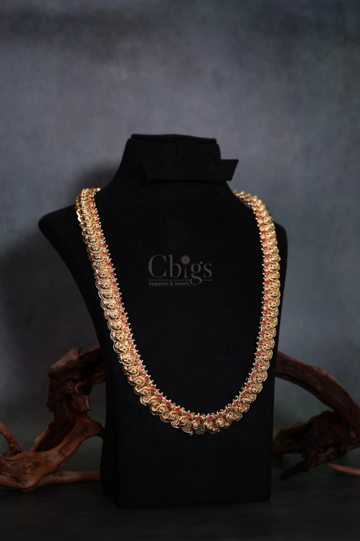 A thick gold-toned chain necklace embellished with red stones, displayed on a black velvet jewelry bust with decorative wood pieces in the background.